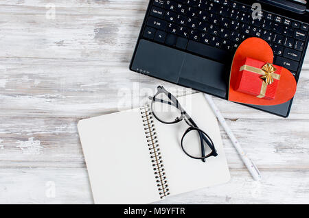 laptop, blank notebook with pen, glasses and gift with heart on wooden table over  workplace. Top view. The concept of love and Valentine's day in off Stock Photo