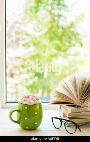 Coffee cup with cream and marshmallows, open book and glasses on window with bokeh. Reading and breakfast. Concept cozy home sweet home Stock Photo
