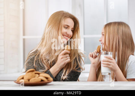 Photo of two laughing sisters - younger and oder. Both girls have blonde straight hair. They are eating delicious sweet cookies from plate and drinking milk near white kitchen's table. Stock Photo