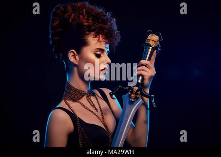 Portrait of young model with make up holding steel sword with black skull. Warrior wearing stylish hairdress with little curles. Girl is wearing black top with thin chains. Dark blue background. Stock Photo