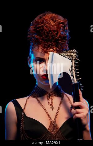 Portrait of girl keeping original sharp metallic axe with thorns. Confident model wearing in black with opened shoulders, with make up and curly hairdress. Chains with crosses on neck of female. Stock Photo