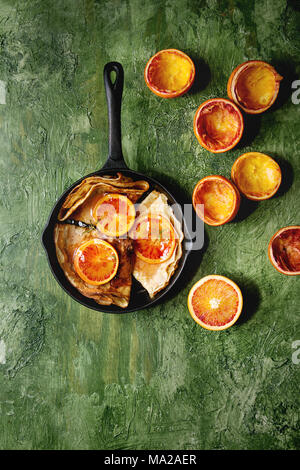 Homemade crepes pancakes served in cast-iron pan with bloody oranges and rosemary syrup with sliced sicilian red oranges over green texture background Stock Photo