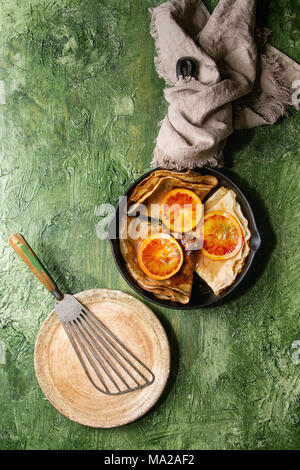 Homemade crepes pancakes served in cast-iron pan with bloody oranges and rosemary syrup with sliced sicilian red oranges and empty plate over green te Stock Photo