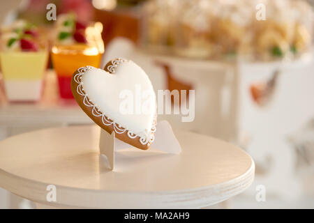 Close-up of a delicious heart-shaped glazed cookie standing on the wooden stand near the candybar with different desserts like yellow cupcakes and red jellies. festive choice gor wedding celebration. Stock Photo