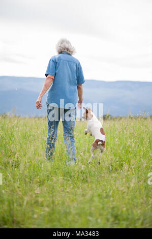 A candid portrait of a senior female walking in a field of tall grass with her small dog jumping alongside.