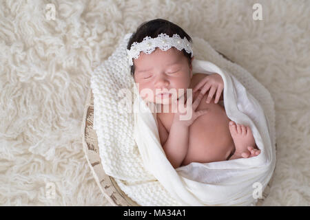 A 12 day old newborn baby girl sleeping in a little, wooden bowl. She is wearing a white, lace and pearl headband. Shot in the studio on a white floak Stock Photo