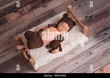 Two week old newborn baby boy wearing a brown, crocheted, bear bonnet. He is sleeping on a tiny, wooden bed and cuddling a stuffed animal. Stock Photo