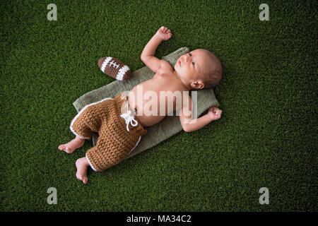 A two week old, alert, newborn baby boy lying on grass turf with a crocheted American football. Stock Photo
