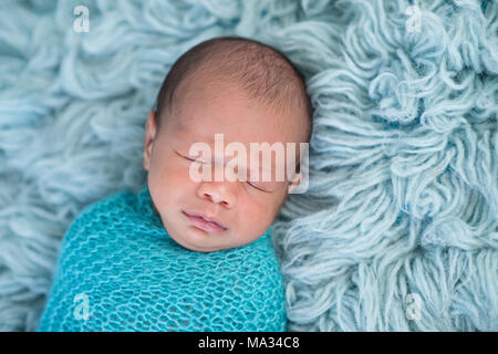 Head shot of a sleeping, two week old newborn baby boy swaddled in a blue wrap. Shot in the studio on a flokati rug. Stock Photo