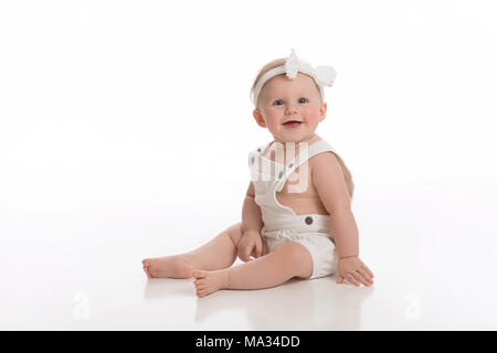 A smiling seven month old baby girl wearing white overalls. Shot in the studio on a white, seamless backdrop. Stock Photo
