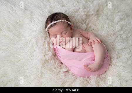 Sleeping, ten day old newborn baby girl swaddled in a light pink wrap. Shot in the studio on a white sheepskin rug. Stock Photo