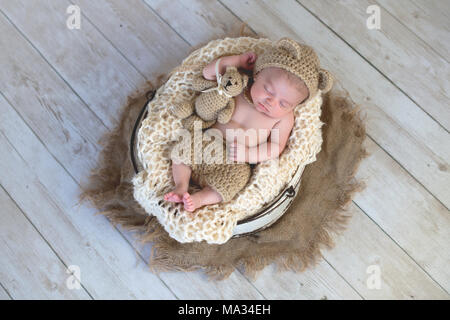 Six week old baby boy wearing a beige, crocheted, bear bonnet. He is sleeping with a matching Teddy Bear. Shot in the studio on a light wood backgroun Stock Photo