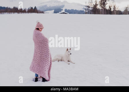 young woman stands wrapped in a pink blanket in a winter landscape,dog lies next to her in the snow, Stock Photo