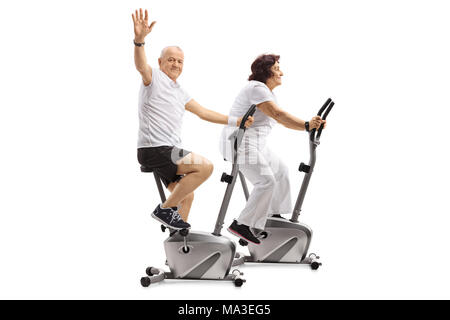 Elderly man and an elderly woman riding exercise bikes with the man waving at the camera isolated on white background Stock Photo