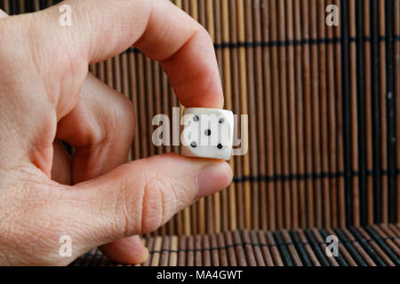 One white plastic dices in man's hand on brown wooden table background. Six sides cube with black dots. Number 5 Stock Photo