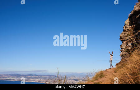 African man standing on a mountain outdoors with his arms and hands wide open facing a magnificent view and clear blue sky over an amazing city scape. Stock Photo