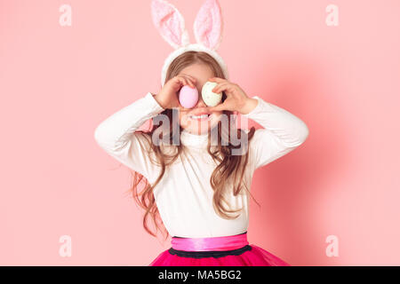 Little child girl wearing bunny ears studio isolated on pink background easter celebration covering eyes with colorful eggs smiling joyful Stock Photo