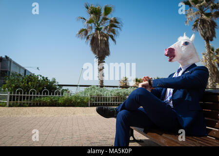Young man in elegant suit sits on the bench on the city street. Unusual guy in funny mask have a break in park. Unicorn is enjoying outdoors Stock Photo