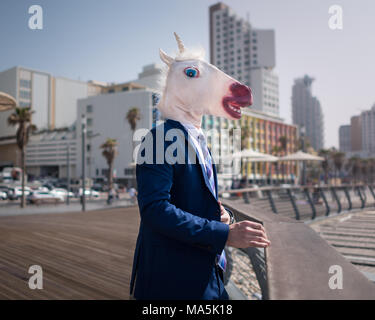 Unusual manager in funny mask and suit stands on city promenade. Young stylish man on the background of cityscape. Unicorn relaxes in warm sunny day Stock Photo