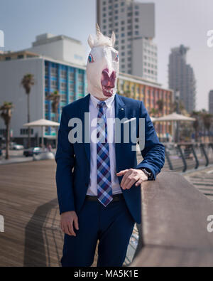 Young elegant man in funny mask and suit stands on city promenade. Unusual manager on background of cityscape. Unicorn relaxes in warm sunny day Stock Photo