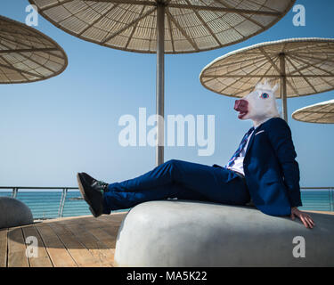 Young man in suit sits under umbrellas on city waterfront. Unusual manager in funny mask have a break. Unicorn is enjoying warm weather Stock Photo