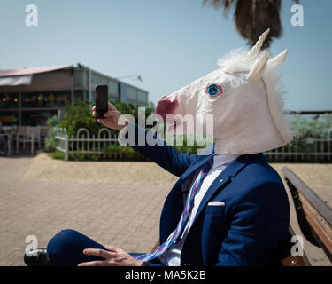 Young unusual man in comical mask and elegant suit makes a photo by phone. Funny unicorn is enjoying warm summer day. Strange guy sits on bench Stock Photo