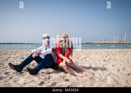 Unusual couple sits on beach near sea. Elegant man in funny mask and sunglasses relaxes with young woman in bright red dress. Freaky unicorn in suit Stock Photo