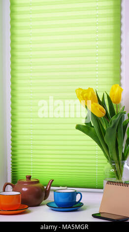 On windowsill there are yellow tulips, multi-colored tea cups, teapot, empty notebook for notes and cell phone. Stock Photo