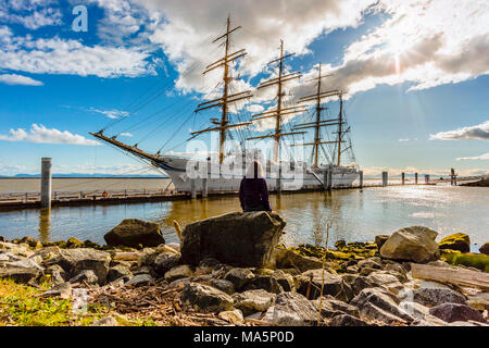 The girl sits on a rock and looks at the big white sailing ship moored to the pier, the blue sky and the rays of the sun making their way through the  Stock Photo