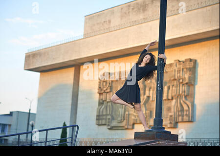 Ivano-Frankivsk, Ukraine - 1 June 2015 : Joung girl is dancing ballet on the street near lamp pillar. Intresting building's facade on background. Model wearing balck dress and having long curly hair. Stock Photo