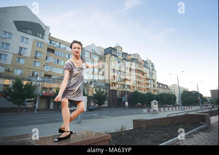 Ivano-Frankivsk, Ukraine - 1 June 2015 : fancy young lady is dancing on city building's facades and blue clear mornig's sky. Woman wearing grey velor dress, necklace and black shoes. Stock Photo