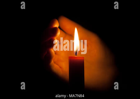 A burning candle at night, protected by the hand of a woman. Candle flame glowing on a dark background with free space for text. Close up, shallow dep Stock Photo