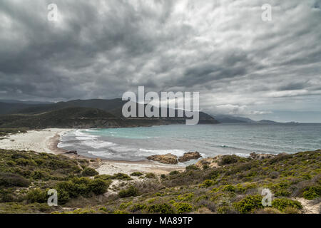 Rocky coastline of Desert des Agriates and Ostriconi beach in Corsica with Ile Rousse in the distance under grey stormy skies and maquis in the foregr Stock Photo