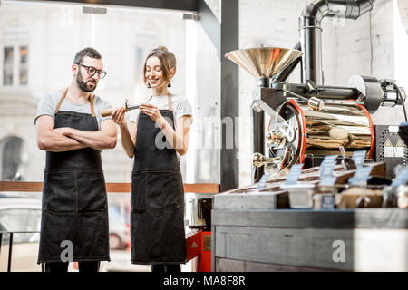 Couple of baristas in uniform checking the quality of roasted coffee beans standing near the roaster machine Stock Photo