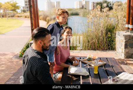 Cheerful couple with their child having a day out. Family sitting in a park with snacks for picnic. Stock Photo
