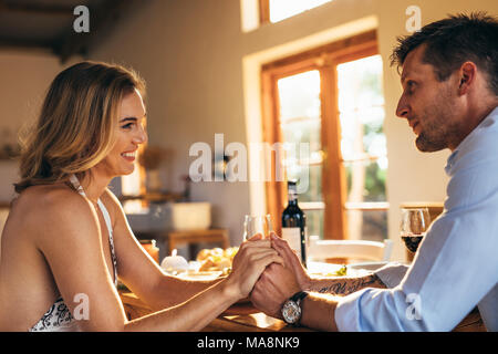 Loving young couple sitting at dining table holding each other's hands. Man and woman having a romantic dinner at home. Stock Photo