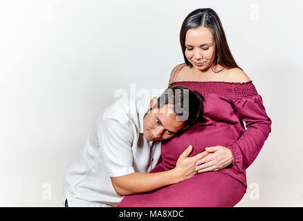 Attractive young pregnant woman wearing a burgandy sleeveless dress  sitting while looking down at her husband who is listening to the baby with his e Stock Photo