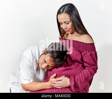 Attractive young pregnant woman wearing a burgandy sleeveless dress  sitting while looking down at her husband who is listening to the baby with his e Stock Photo