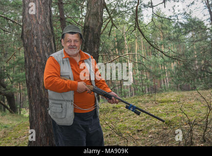 Portrait of positive and smiling senior ranger with rifle in pine forest Stock Photo