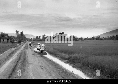 Black and white photograph showing several people, wearing conical woven hats (non la) and carrying baskets hanging from bamboo poles, walking down a dirt, country road, with a house at left and rice paddies at right, photographed in Vietnam during the Vietnam War (1955-1975), 1968. () Stock Photo