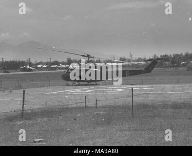 Black and white photograph showing a helicopter docked on an airstrip, with trees and buildings, likely a US Marine Corps military camp, in the background, photographed in Vietnam during the Vietnam War (1955-1975), 1968. ()