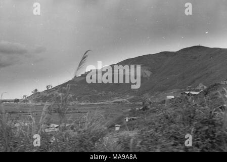 Black and white photograph, shot from the vantage of a hill, showing the Vietnamese countryside, with grass-covered fields or rice paddies, the roofs of houses or barracks, and a large hill or mountain, capped with an observation tower, in the background, photographed in Vietnam during the Vietnam War (1955-1975), 1968. () Stock Photo