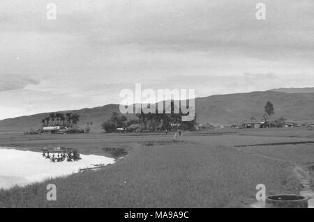 Black and white photograph showing a country landscape, with a lake and rice paddies in the foreground, houses and trees at midground, and a hill in the background, photographed in Vietnam during the Vietnam War (1955-1975), 1968. () Stock Photo