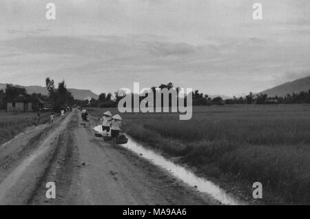 Black and white photograph showing several people, wearing conical woven hats (non la) and carrying baskets hanging from bamboo poles, walking down a dirt, country road, with a house at left and rice paddies at right, photographed in Vietnam during the Vietnam War (1955-1975), 1971. () Stock Photo