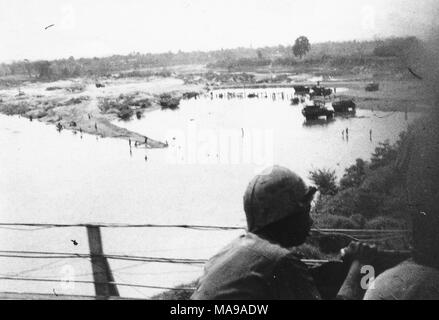 Black and white photograph, showing the back of a helmeted soldier, presumably riding in a truck over a bridge, with a body of water, boats, flat land, and trees in the background, photographed in Vietnam during the Vietnam War (1955-1975), 1971. () Stock Photo