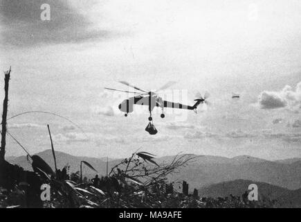 Black and white photograph showing a helicopter, in mid-flight, carrying large bags, and silhouetted in profile against a cloudy sky, with mountains in the background, and foliage and a fence in the foreground, photographed in Vietnam during the Vietnam War (1955-1975), 1971. ()