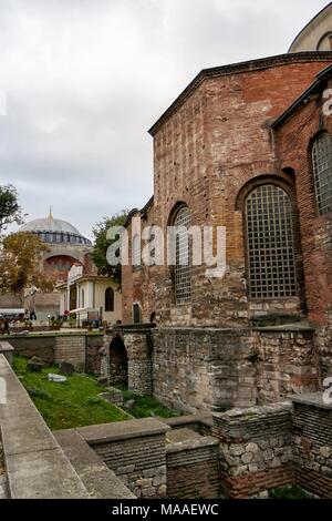 Exterior view, showing the Hagia Sophia Mosque in the background at left, and the facade, with arched windows, of the Romanesque, stone and brick built Aya Irini (Hagia Irene), a former Greek Eastern Orthodox church, now used as a museum and concert hall, located in the outer courtyard of Topkapi Palace in Istanbul, Turkey, November 16, 2017. () Stock Photo