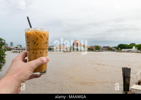 https://l450v.alamy.com/450v/maamga/a-male-hand-holds-out-a-clear-glass-of-thai-iced-coffee-with-ice-cubes-and-plastic-straw-with-a-buddhist-temple-and-large-river-in-the-background-ban-maamga.jpg