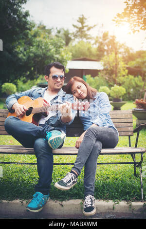 Young couple in love playing acoustic guitar in the park while sitting on the bench together. Stock Photo