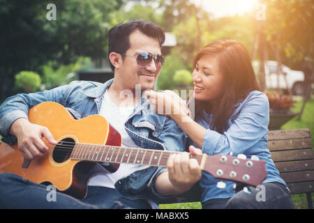 Young couple in love playing acoustic guitar in the park while sitting on the bench together. Stock Photo